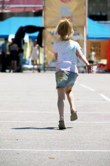 Fototapeta premium A little girl runs and skips across the asphalt towards a some woman during the Holi festival celebration. On the back of her skirt you can see a yellow and blue heart with red spots.