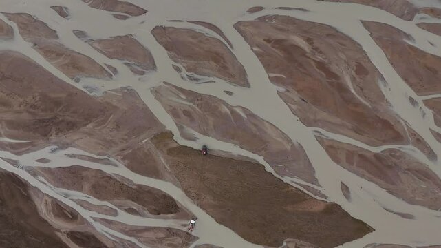 An aerial view shows several vehicles navigating the intricate patterns of a braided river system within a vast, earthy landscape