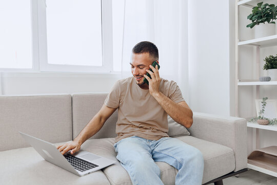 Engaged man making a phone call while working on a laptop, sitting comfortably on a modern couch, showcasing a productive and relaxed home office atmosphere.