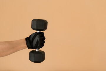 Mans’s hand wearing a black gym glove gripping a heavy hexagonal dumbbell horizontally, isolated against a warm neutral gold-beige background