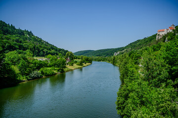 Fototapeta premium Scenic river flowing through lush greenery with a historic castle overlooking the landscape in bright daylight