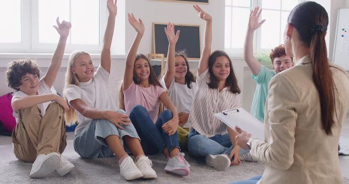 Smiling high school children sitting on carpet in classroom in front of female teacher, raising their hands up to answer question or to vote, studying together in relaxed and cheerful atmosphere.
