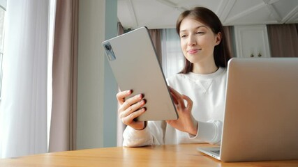 A young woman enjoys online learning on her tablet and laptop in a sunlit, decorated room.