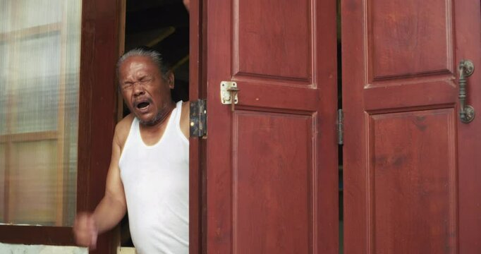 Asian man which is senior, stands at the doorway of his wooden home, quietly opening the door to a new day. His gaze and body language reflect solitude, routine, and a silent resilience.


