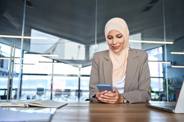 Middle Eastern muslim businesswoman manager ceo using cell phone mobile app. Smiling young indian woman in hijab holding smartphone sitting in office working online at desk on gadget with copy space.