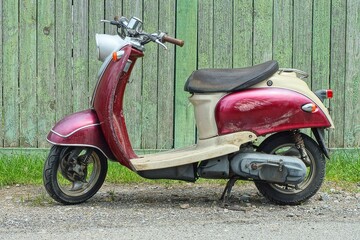 one cherry plastic small modern two wheels  industrial with  black soft saddle with motor scooter stands on the ground near the wooden green fence in the daytime on the street	