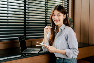 Beautiful Asian woman working in coffee shop. Portrait of freelancer working in cafe using laptop and notebook.