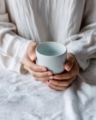 Hands in pale blue sleeves holding ceramic cup filled with water over white linen, soft shadows and tactile textures, peaceful still moment