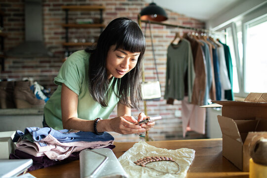 Woman photographing handmade jewelry at home for online store surrounded by clothes and packing materials