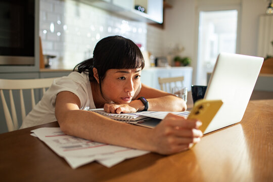 Frustrated woman laying her head on desk while holding phone and looking at laptop at home