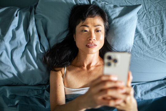 Woman lying in bed using smartphone looking thoughtful while scrolling on screen