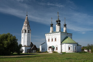 White stone orthodox church with black domes and a tall bell tower standing on a green lawn under a blue sky with some clouds. Church of the Resurrection of Christ, Suzdal