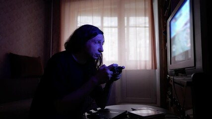 An intensely focused and absorbed mood is captured as a man, illuminated by the blue glow of a screen, plays a video game with a controller in a dimly lit room