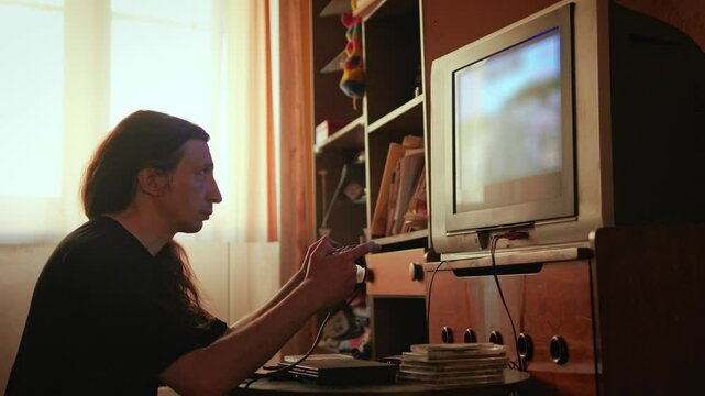 An intensely concentrated and nostalgic mood is captured as a man with long hair plays a video game on an older television set, his profile sharply focused on the screen