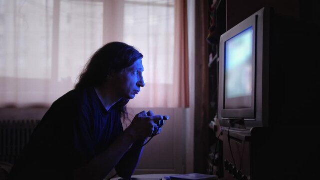 An intensely focused and absorbed mood is captured as a man, illuminated by the blue glow of a screen, plays a video game with a controller in a dimly lit room