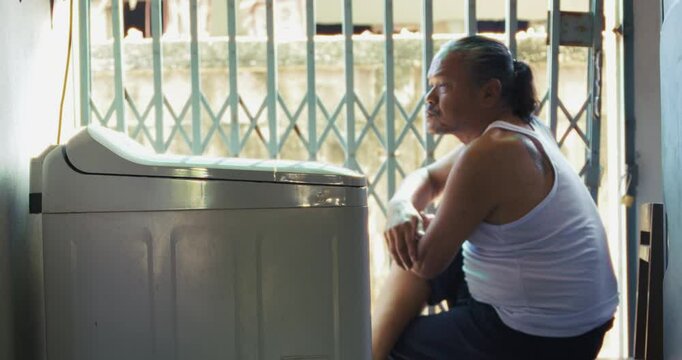 Asian man which is senior, sits beside a washing machine behind his house. Surrounded by concrete and daily items, he does his laundry in silence, his face lost in memory and quiet thought.