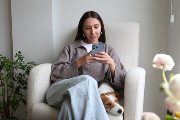 Young woman scrolling the phone at home with her dog. Copy space, background.