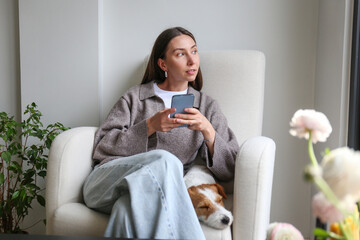 Young woman scrolling the phone at home with her dog. Copy space, background.