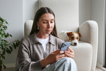Young woman scrolling the phone at home with her dog. Copy space, background.