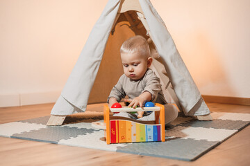 Cheerful 2 year old boy sitting near the wigwam and playing with a wooden toy. Toddler boy in a beige t-shirt sitting in the living room and playing with toys. Child in comfortable home clothes
