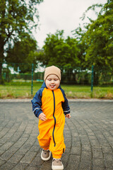 Smiling 2 year old boy in mudproof overalls and boots runs after the rain. Toddler in bright...