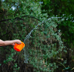 Child playing with water in a garden on a sunny day, creating splashes with an orange bucket