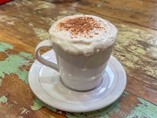 Close up of a cappuccino in a white mug with foam and chocolate sprinkles on a wooden table top