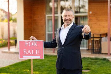 Male real estate agent in suit posing near house sale sign and showing keys, property investment...