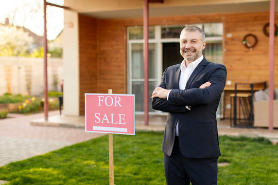 Successful man real estate agent in suit posing near sign for sale outdoors in front of house, smiling at camera with folded arms