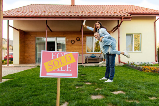 Excited couple new homeowners posing in front of their house, having fun, man piggybacking wife, sold sign on foreground. Property and purchase concept - Powered by Adobe