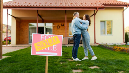 Man and woman new homeowners celebrating purchase, embracing in front of house and board with sold...