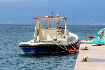 Compact white and blue tugboat with red trim is tied to a concrete pier, floating on calm sea water with red buoys and a distant coastline under a cloudy sky. © hecos