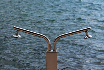 Modern stainless steel beach shower with two angled heads stands on a dock or platform, positioned over rippling blue water, reflecting light and designed for outdoor use at lakes or seaside.