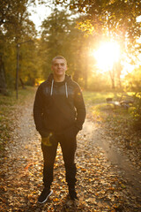 Smiling young man in a black hoodie standing on a forest path in autumn. Warm golden hour sunlight,...