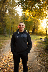 Smiling young man in a black hoodie standing on a forest path in autumn. Warm golden hour sunlight,...