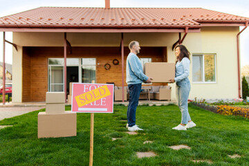 Real estate and property concept. Focus on SOLD sign, couple with boxes in front of their new house on moving day