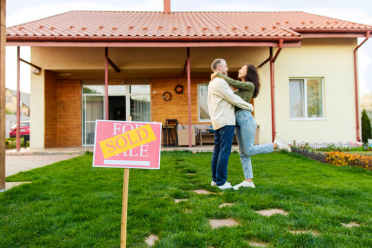 Real estate concept. Focus on sold sign with happy couple embracing on background, celebrating their purchase, posing in front of own house - Powered by Adobe