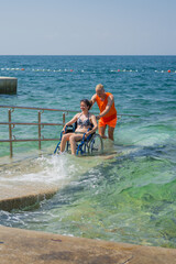 Relaxed smiling woman with disability, exiting the sea water with the help of an assistant, across...