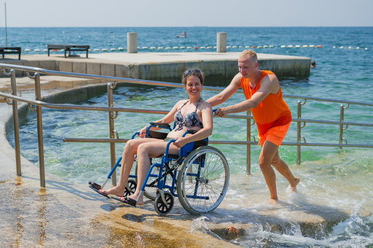 Relaxed woman with disability, exiting the sea water with the help of an assistant, across an accessible wheelchair beach ramp.