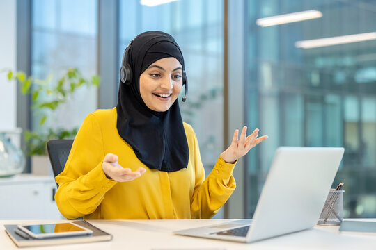 A smiling woman in a yellow shirt and hijab speaks into a headset while looking at her laptop in an office setting.