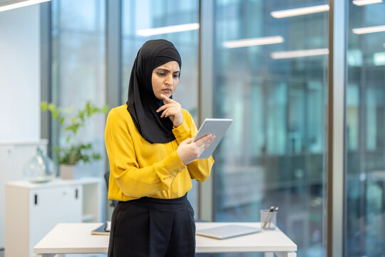 A thoughtful businesswoman in a hijab reviews information on a tablet inside a modern office setting, contemplating her next action. - Powered by Adobe