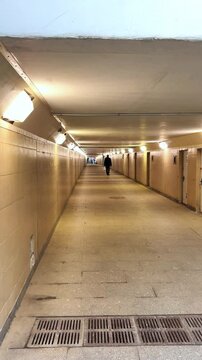 Long, illuminated pedestrian tunnel with smooth walls and floor, featuring a solitary figure walking towards the light at the end, creating a sense of depth and perspective in urban environment