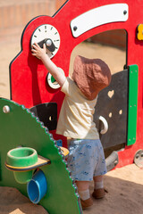Baby boy dressed with summer cloves and hat is playing outside on the street, touching and exploring playground for little ones. The child is reaching up to interact with a red play structure.