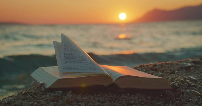 Open book rests on a pebbled beach during sunset with ocean waves in background