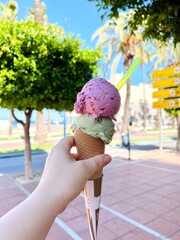 Hand holding a delicious ice cream cone with two colorful scoops, one purple and one green, against a sunny outdoor backdrop featuring palm trees and a clear blue sky, joyful summer experience