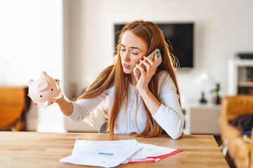 Young woman discussing finances on phone while holding a piggy bank at home during the day