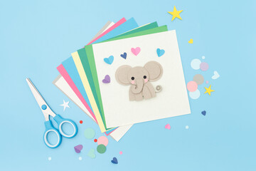 Cute elephant made from colored paper on square white sheet with hearts, stars, and scissors on blue background. flat lay with craft supplies. Animal paper craft for kids. copy space,