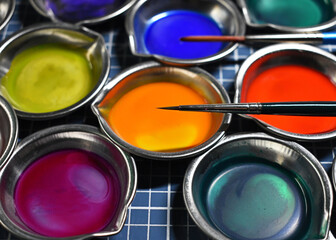 Colorful liquid paints and fine artist paintbrush in small metal dishes for creativity hobby studio work close up