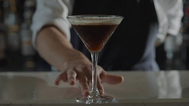 A bartender is reaching out to shake a drink. The bartender is wearing a black apron and a white shirt. The image has a casual and friendly vibe