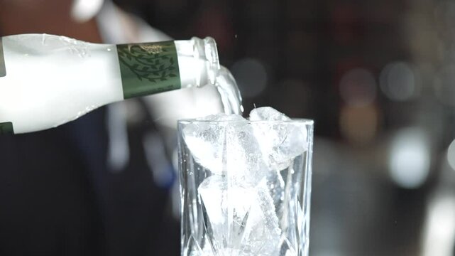 A person is pouring a drink into a glass with ice cubes. The drink is likely a cocktail, as the bottle is labeled with a wine logo